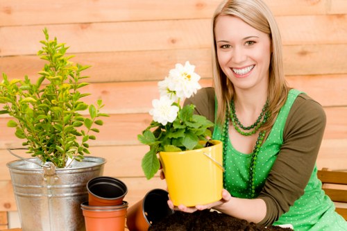 The image shows a young woman with blonde hair smiling and sitting outdoors in a garden area, holding a yellow flower pot with white flowering plants. In front of her on a wooden table are several empty plant pots of varying sizes, including black, terracotta, and smaller black pots. To her left, there is a larger metallic pot containing a green shrub with dense, bright green leaves. The background features a wooden fence or wall, suggesting a well-maintained garden or patio space. The scene is set during daylight, with natural lighting highlighting the lush greenery and vibrant colors of the flower pots, indicating an environment suitable for gardening and outdoor plant care. The woman is dressed in a green top with a polka-dot pattern, complementing the gardening theme, and her cheerful expression reflects a positive connection to outdoor gardening activities, aligning with the services offered by Hedge Trimming Crouch End for garden maintenance and plant care.