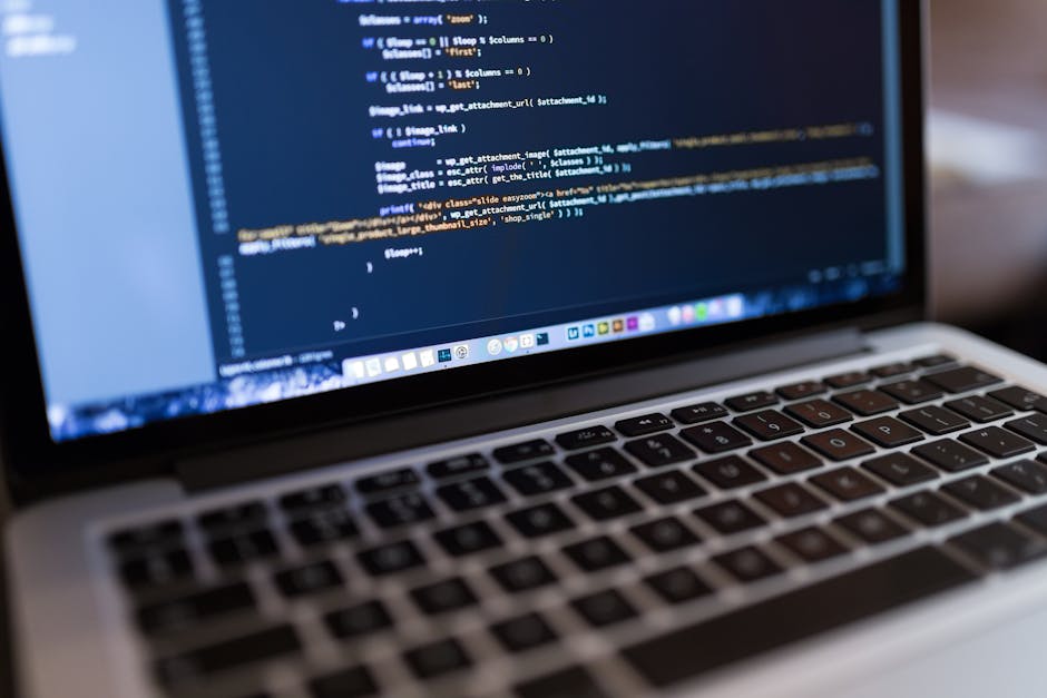 Close-up of a laptop keyboard and screen displaying lines of programming code, with the code highlighted in various colors such as white, blue, and orange. The scene is indoors, with the focus on the