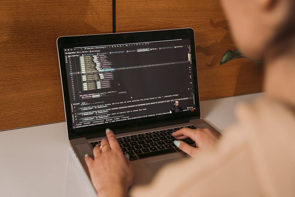 A person with light skin and blonde hair, wearing a beige top, is seated at a white table, working on a laptop with a dark-themed programming or coding interface displayed on the screen. The laptop is