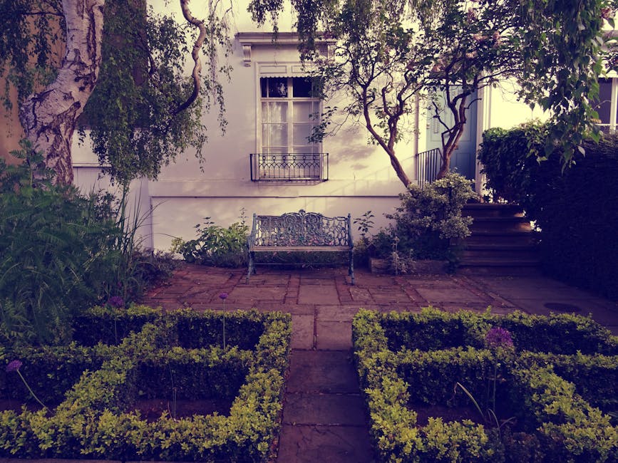 A neatly maintained garden features a paved stone patio area with a decorative wrought iron bench situated against a white exterior wall of a house. The garden is framed by lush greenery, including fl
