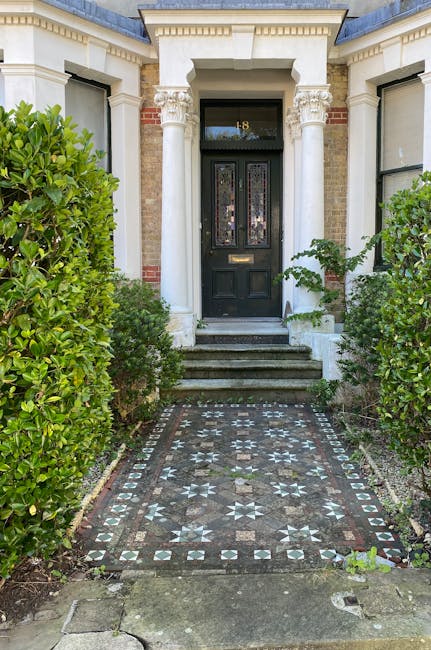 A neatly maintained garden features a paved stone patio area with a decorative wrought iron bench situated against a white exterior wall of a house. The garden is framed by lush greenery, including fl